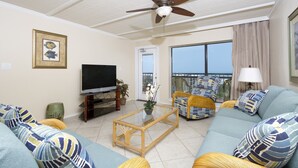 Un salón con ventilador de techo, televisor de pantalla plana, muebles de mimbre y un amplio ventanal con vistas.