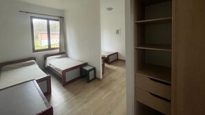 A room with two single beds, a window with a view of a house, a wooden shelf unit, and a small table.