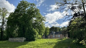 A large, historic building with multiple towers and a stone wall, surrounded by lush greenery and trees.