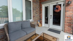 Eine Rattan-Couch auf einer Veranda mit einer Ziegelwand, einer Tür mit einem Kranz und einer Lampe.