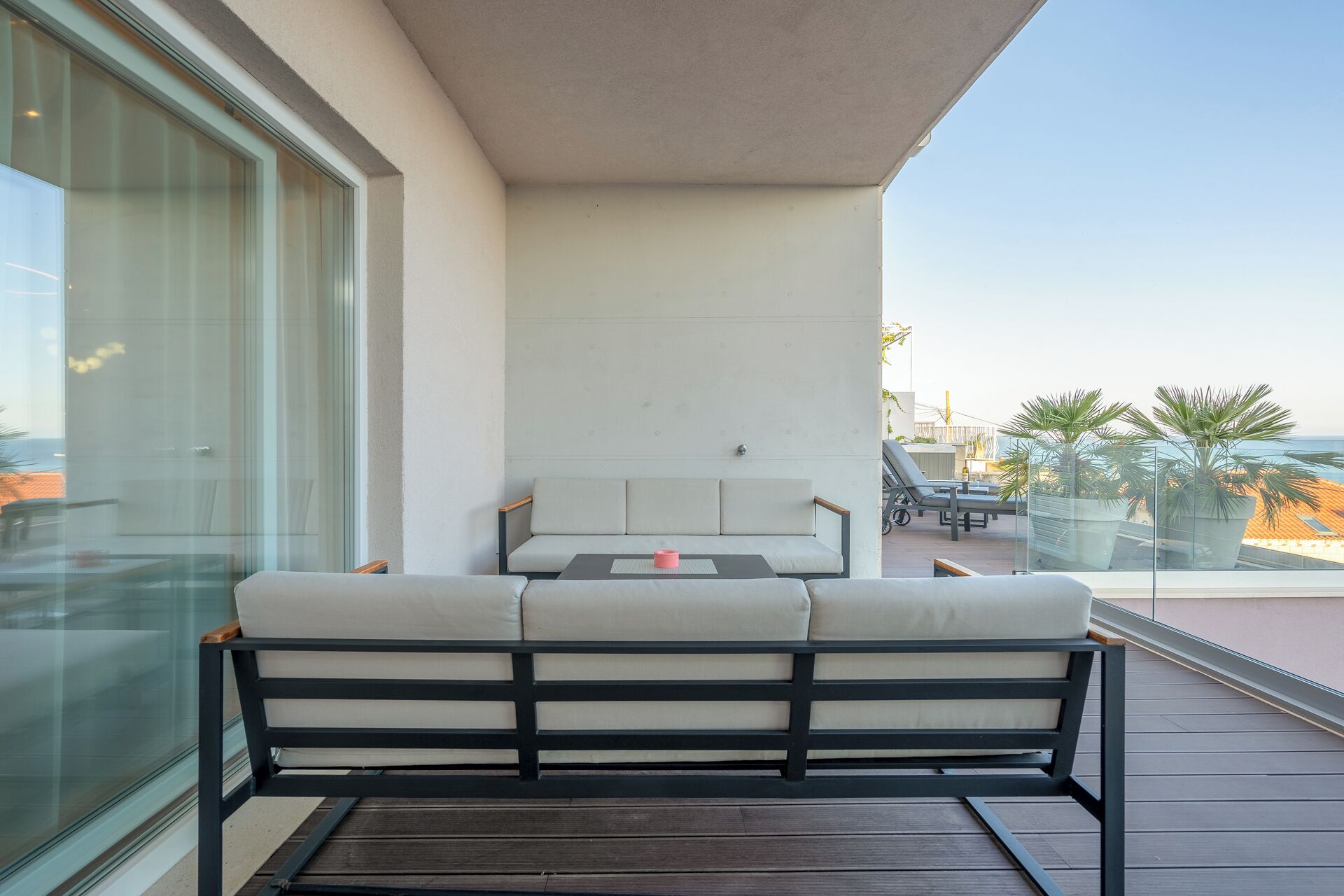 A modern outdoor seating area with a sofa and table, overlooking a beach and palm trees.