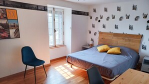 Un dormitorio con cabecera de madera, cama con ropa de cama azul y dos almohadas amarillas, un escritorio de madera, una silla azul y una ventana con persianas.