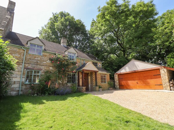 A stone house with a garage, surrounded by a well-maintained lawn and trees. - 1 The Lane (Moreton-in-Marsh)