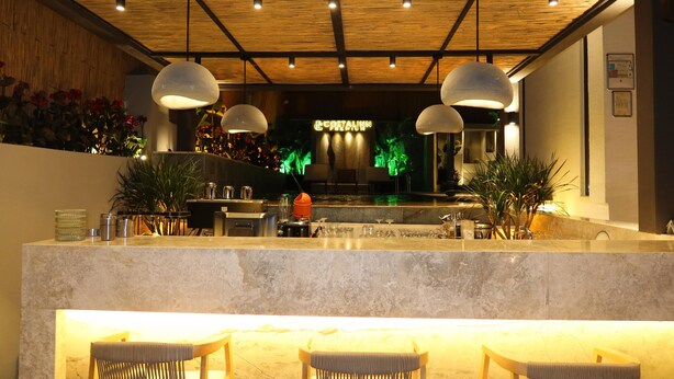 Una zona de bar con mostrador de mármol, luces colgantes tipo lámpara y una recepción con un letrero de neón.