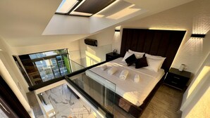 Un dormitorio moderno con balcón de cristal, una cama grande con almohadas, una mesita de noche y un velador.
