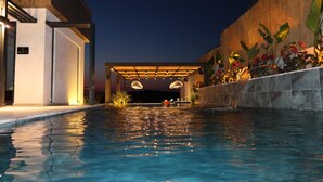 Piscina con un porche de madera cubierto, rodeada por un muro de piedra y plantas tropicales.