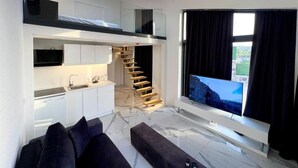 Un apartamento moderno con un dormitorio en entrepiso, una zona de cocina, una escalera y una pantalla de televisión grande.