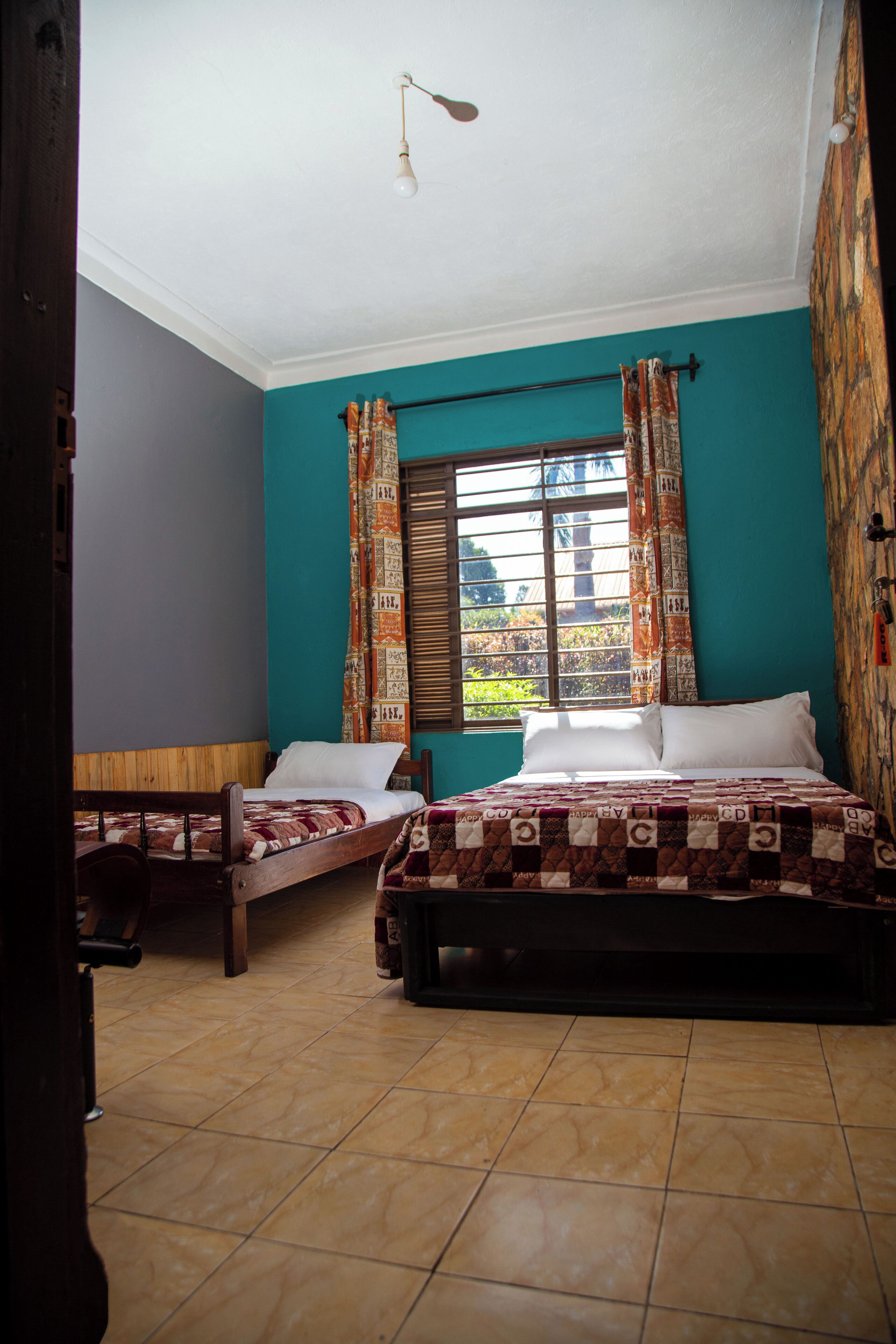 A room with two beds, a window with curtains, a ceiling light, and a tiled floor.