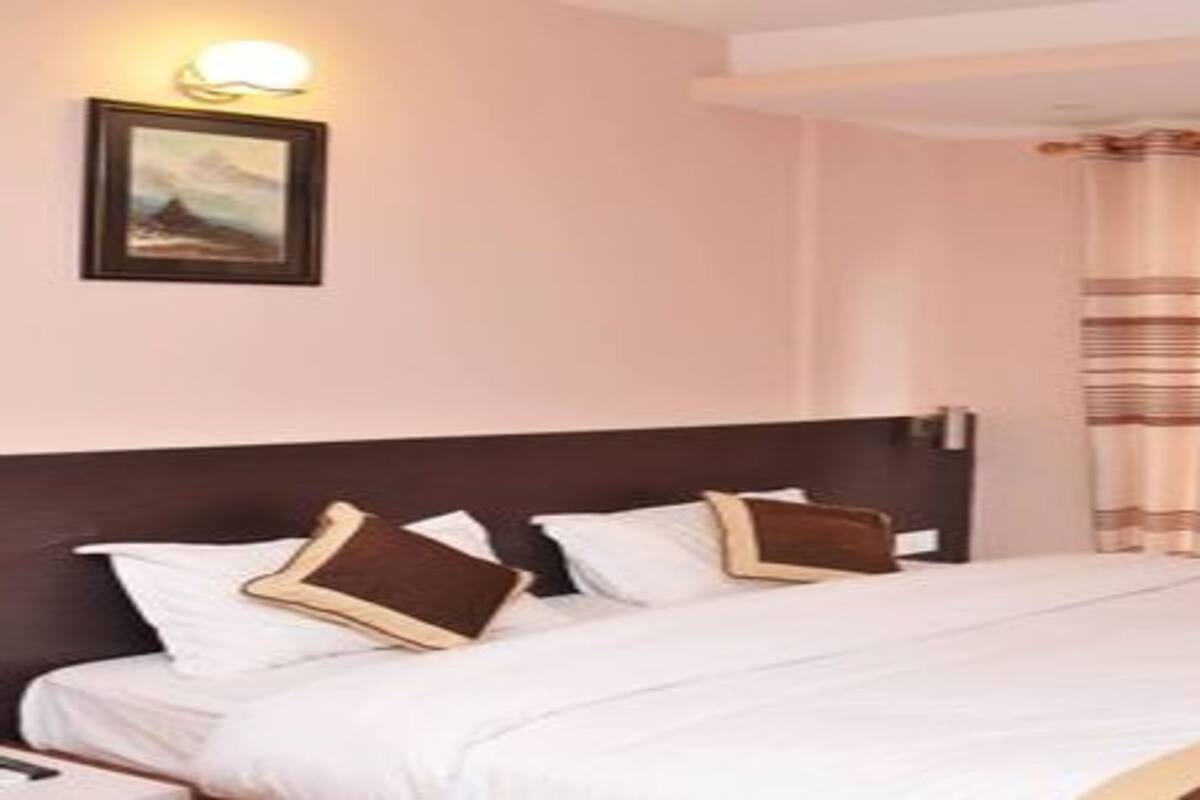 Habitación de hotel con cama, almohadas, cabecera, luz empotrada en la pared y ventana con cortinas.