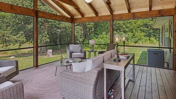 A covered outdoor seating area with wicker furniture, a ceiling fan, and large windows offering a view of the surrounding greenery. - Rocky Flats Haven (Cosby)