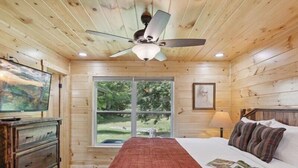 A wooden cabin bedroom with a bed, dresser, ceiling fan, and a window with a view of greenery. - Rocky Flats Haven (Cosby)