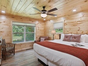 A wooden cabin bedroom with a bed, a chair, a ceiling fan, and a window with a view of greenery.