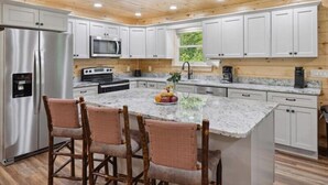 A kitchen with white cabinets, a stainless steel refrigerator, a microwave, and a stove. There are three wicker chairs at the island. - Rocky Flats Haven (Cosby)