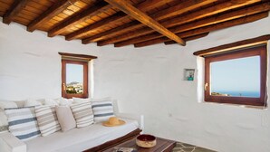 A cozy room with a wooden ceiling, a white sofa with striped pillows, a small table with a bowl and a mug, and a window with a view of the sea.