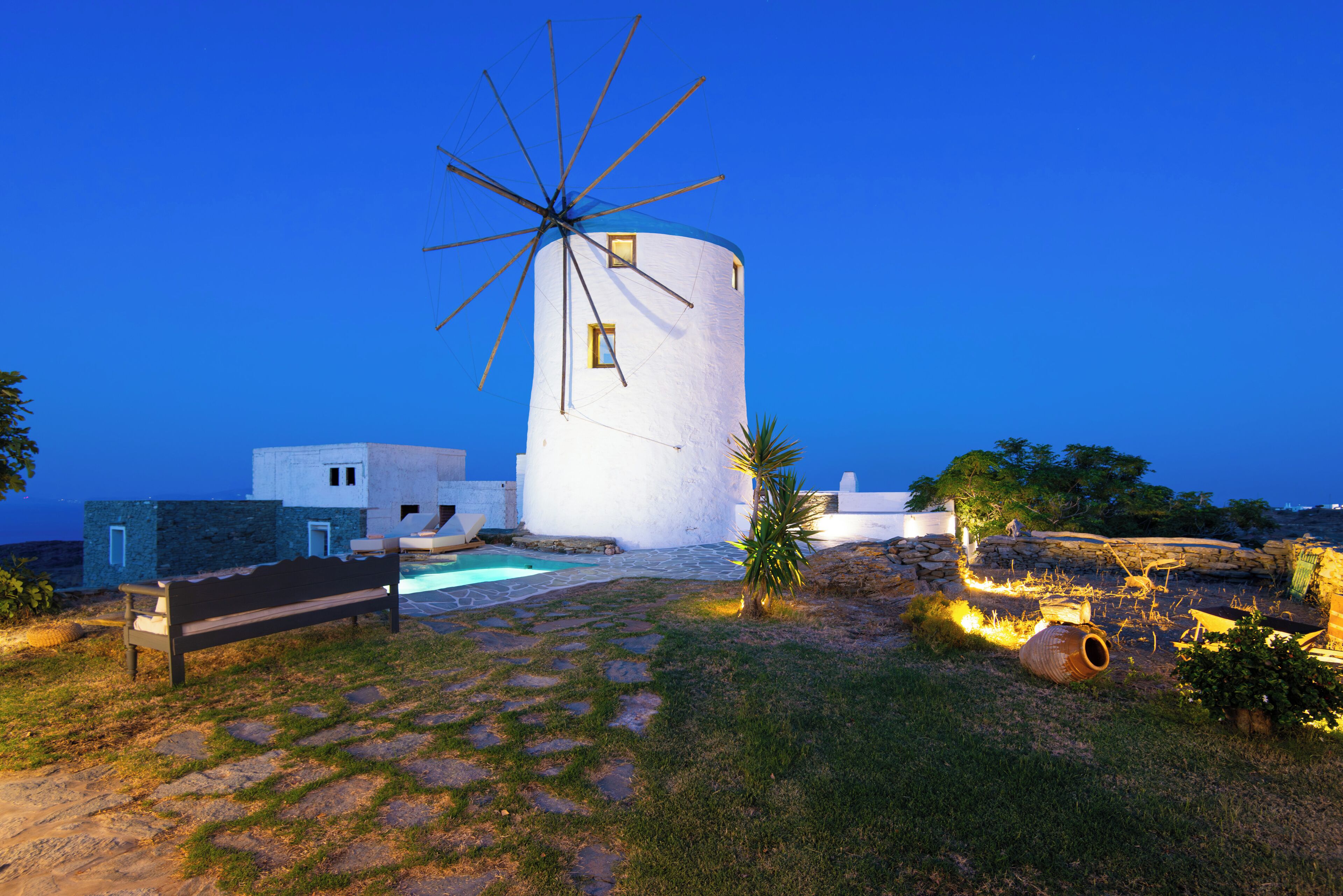 Un moulin à vent aux murs blancs, avec un ciel bleu en arrière-plan. On aperçoit un banc et une petite piscine au premier plan.