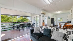 A modern living room with a wooden deck, a sofa with patterned cushions, a dining area with a table and chairs, a kitchen with a refrigerator and microwave, and large windows offering a view of greenery. - The Cottage - 16 Clarke Street (Bargara)