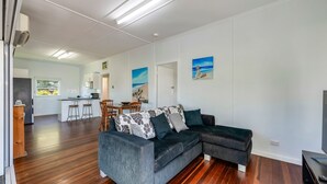A modern living room with a grey sectional sofa, wooden flooring, and a dining area with a kitchen in the background. - The Cottage - 16 Clarke Street (Bargara)