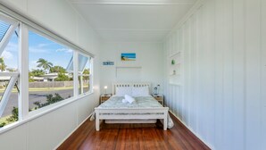 A bedroom with a bed, a nightstand, a wall-mounted shelf, and a window with a view of the street and palm trees. - The Cottage - 16 Clarke Street (Bargara)