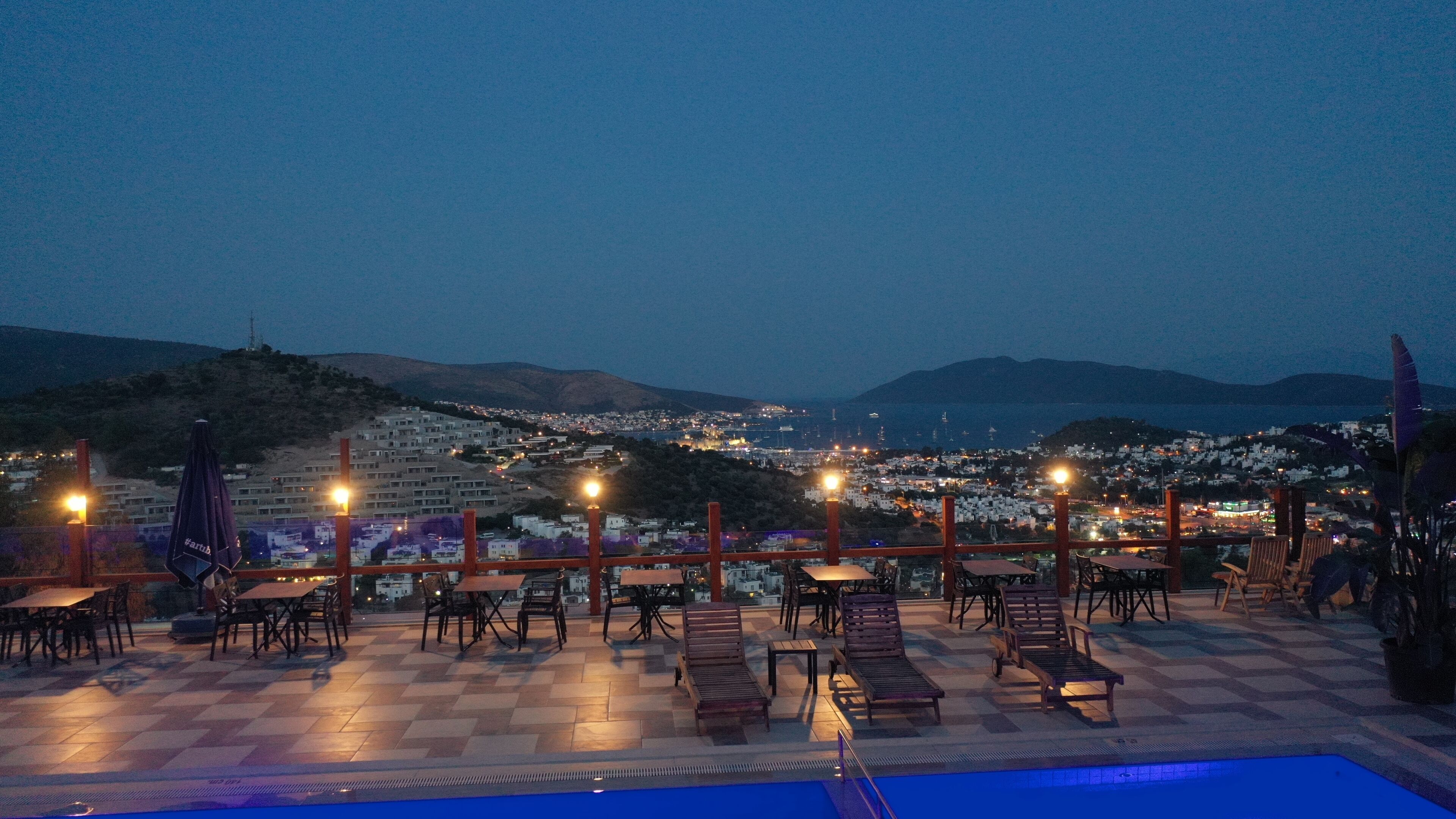 A terrace with outdoor seating and a pool, overlooking a cityscape at dusk.