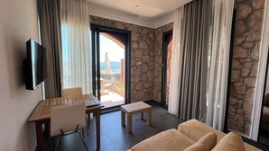 Habitación de hotel con pared de piedra, un televisor montado en la pared, un escritorio con una silla, un sofá y vistas al exterior a través de una puerta de vidrio.