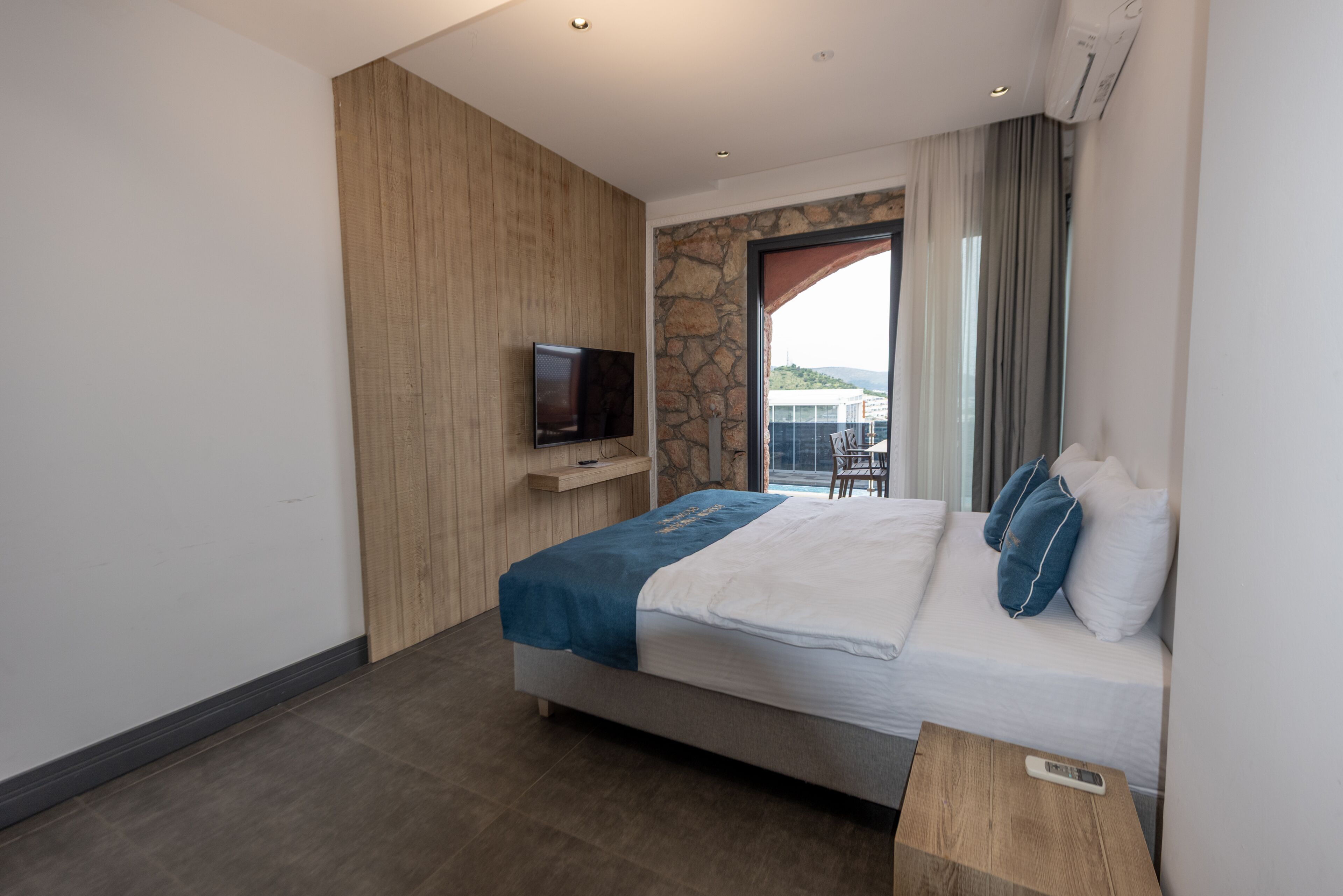 Una habitación de hotel moderna con cama, televisor, pared decorativa de piedra y balcón con vistas.