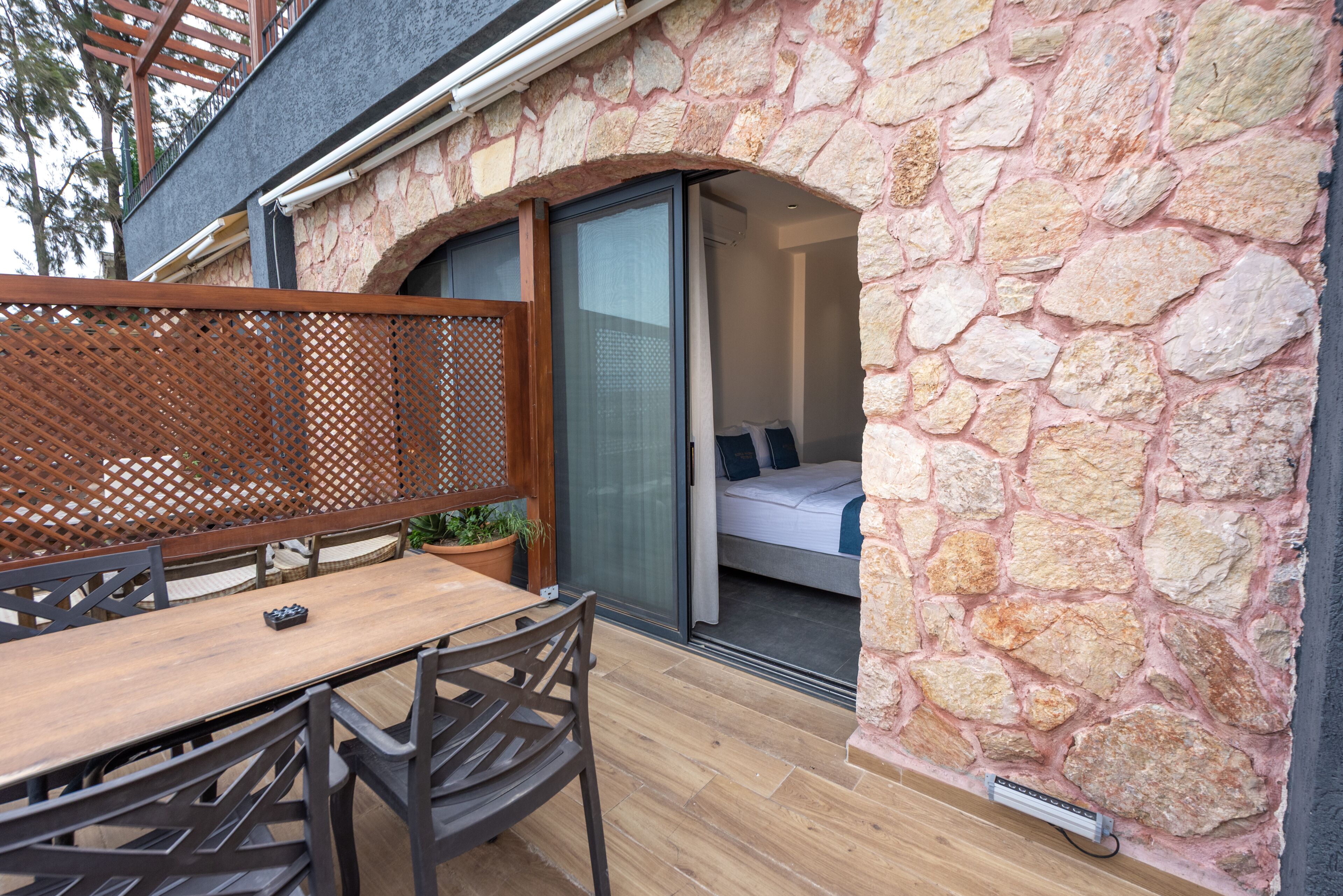 A stone exterior wall, a wooden table with chairs, and a sliding glass door leading to a bedroom with a bed and pillows.