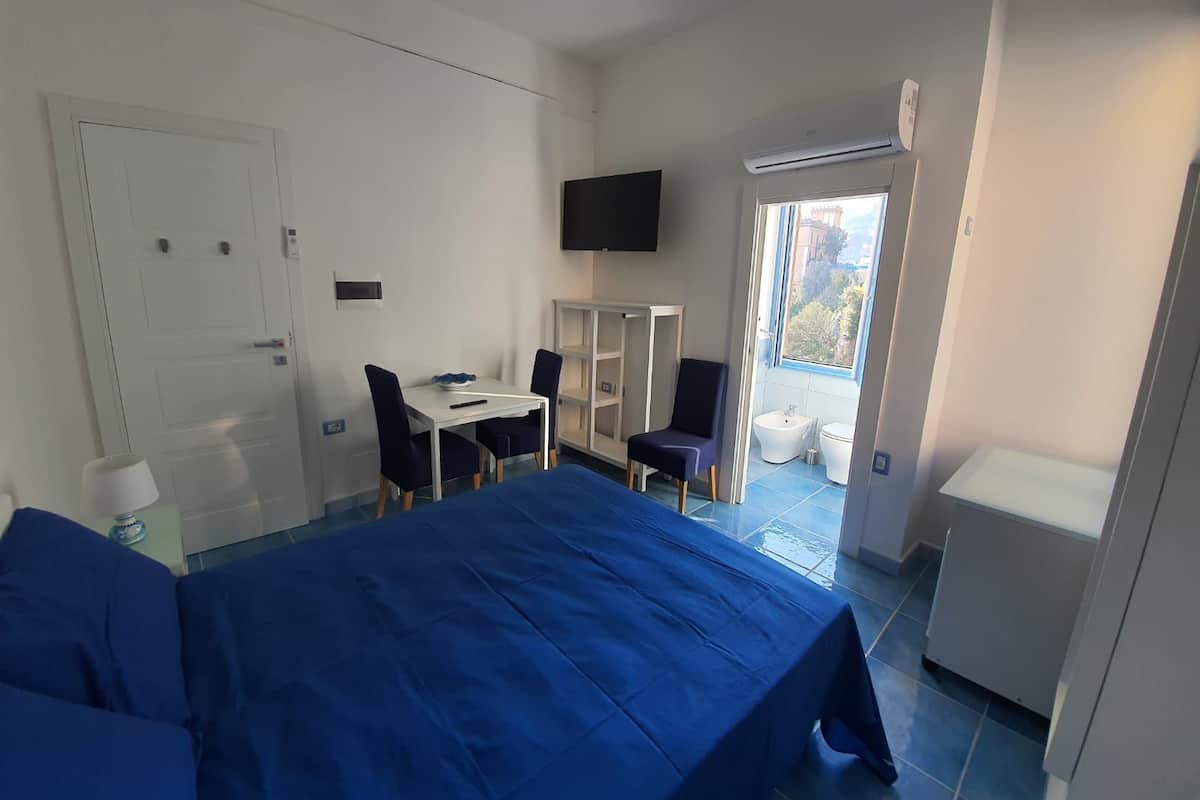 Quarto de hotel com cama, escrivaninha com cadeiras, televisão, janela e banheiro.