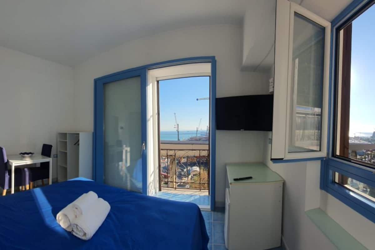 Quarto de hotel com cama, escrivaninha, cadeira, mesinha, televisão e janela com vista para o mar e um edifício.