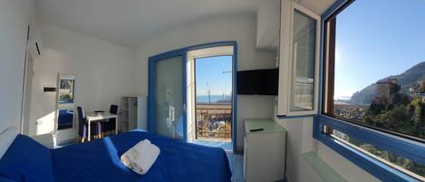 Habitación de hotel con cama, escritorio, silla, mesita, televisión y ventana con vista al mar y un edificio.
