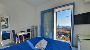 A hotel room with a bed, a desk, a chair, a TV, and a balcony with a view of the sea and cranes. - B&B Acquamarine (Salerno)