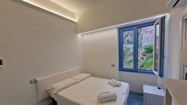 A small, modern bedroom with a single bed, a small table, and a window with a view of greenery. - B&B Acquamarine (Salerno)