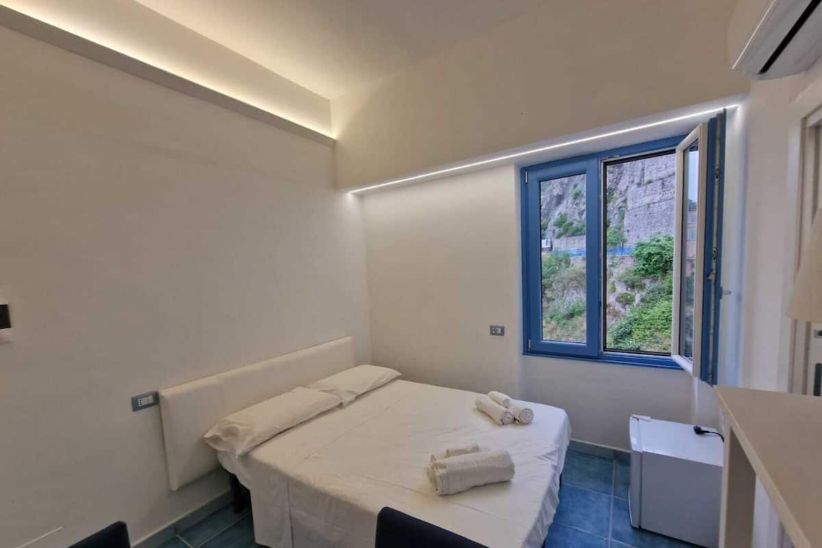 Quarto pequeno e moderno com uma cama de solteiro, uma mesinha e uma janela com vista para vegetação.