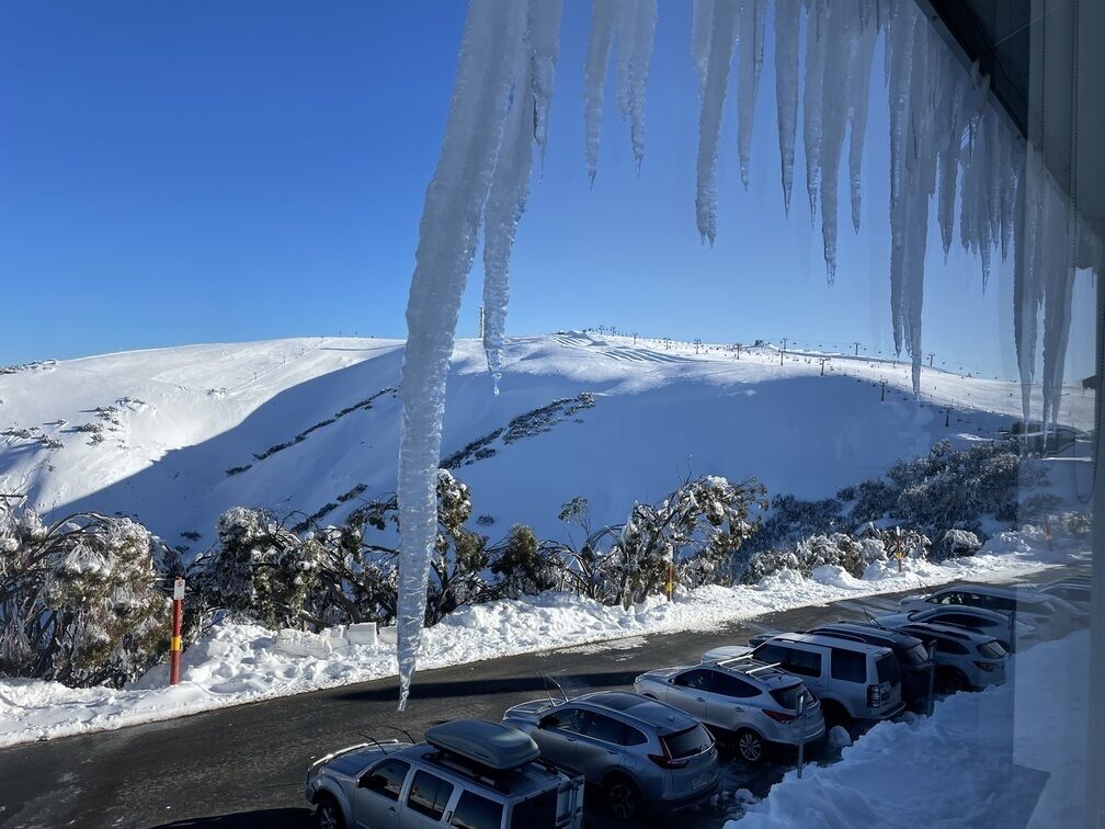 Ein Parkplatz mit Autos und eine verschneite Landschaft, von denen Eiszapfen an der Dachrinne hängen.