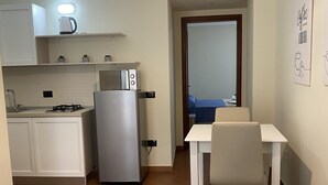 A compact room with a kitchenette, a bed with a blue quilt, a white chair, and a hanging light fixture. - 2 Bedroom Apartment in Realmonte (Realmonte)