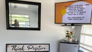A room with a framed motivational sign, a mirror, a small table with a potted plant, and a chair.