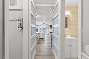 A narrow hallway with a bed, a table, and a chair. There is a window with curtains and a ceiling fan. - Top of the Gulf #120- Free Beach Chairs- Shore-Side Bungalow | Perfect little family getaway! (Panama City Beach)