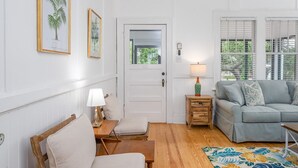 A living room with a grey sofa, a wooden side table with a lamp, a small wooden table, and a floral rug. - Quaint and Cozy Beachside Cottage w/ Screened-In Porch by Tybee Cottages (Tybee Island)