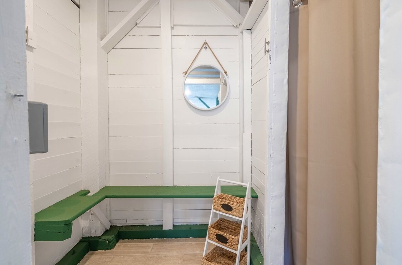 Une petite pièce étroite aux murs blancs et sur laquelle est posé un banc vert. Un miroir rond est accroché au mur, et une échelle blanche, sur laquelle sont fixées des corbeilles, est appuyée contre le mur.