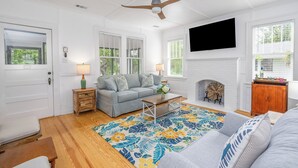 A living room with a grey sofa, wooden coffee table, and a colorful floral rug. There is a ceiling fan, a fireplace, and a TV mounted on the wall. - Quaint and Cozy Beachside Cottage w/ Screened-In Porch by Tybee Cottages (Tybee Island)
