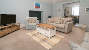 A modern living room with a flat-screen TV, two sofas, a coffee table, and a kitchen area in the background. - The Garden Flat (Bridport)