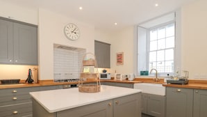 A modern kitchen with grey cabinets, a white countertop, a three-tiered wicker basket, a clock, and a window providing natural light. - The Garden Flat (Bridport)