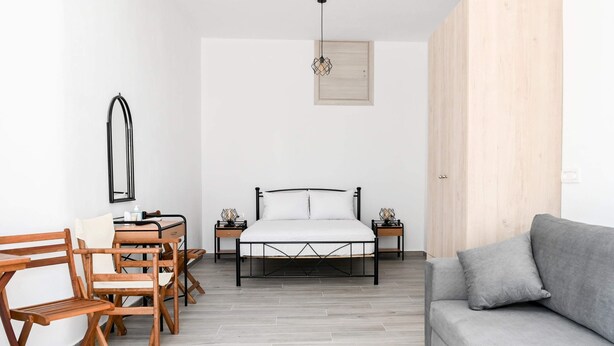 A bedroom with a bed, a sofa, a desk with chairs, a mirror, and a hanging light fixture. - Experience Dreamy Santorini Gorgeous Std (Santorini)