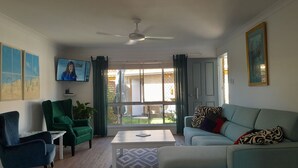A living room with a sofa, armchair, coffee table, and a TV. There are windows with curtains, a ceiling fan, and artwork on the walls. - 2BR villa in quiet cul-de-sac with BBQ, herb garden, games, parking, (Evans Head)