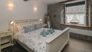 A bedroom with a bed, bedside lamps, a stone wall, a window with a hanging chair, and a wooden ceiling beam. - Teal House (Lyme Regis)