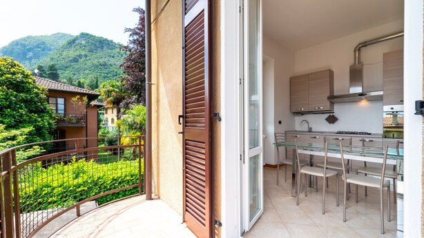 A modern kitchen with a glass dining table and chairs, a balcony with a metal railing, and a view of a lush green landscape. - Bright and Cozy in Menaggio by Wonderful Italy (Menaggio)