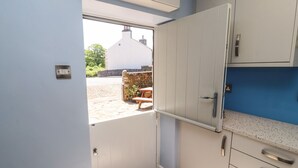 A compact kitchen with white cabinets, a blue countertop, and a door leading to an outdoor area with a stone wall and a bench. - Rose Cottage (Settle)