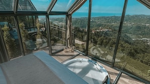 A modern room with a large bed, a hot tub, and a panoramic view of a mountainous landscape through large windows.