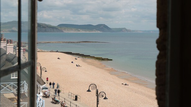 Une plage où l’on peut voir des gens se prélasser au soleil et se promener, une jetée qui s’avance dans l’eau et, au loin, des collines.
