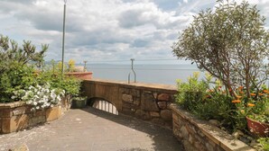 Un chemin de pierre mène à un mur de pierre qui surplombe une étendue d’eau, et des plantes et des fleurs en pot bordent le chemin.