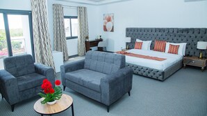 A modern hotel room with a large bed, two armchairs, a small table with flowers, and a view of greenery through the windows.